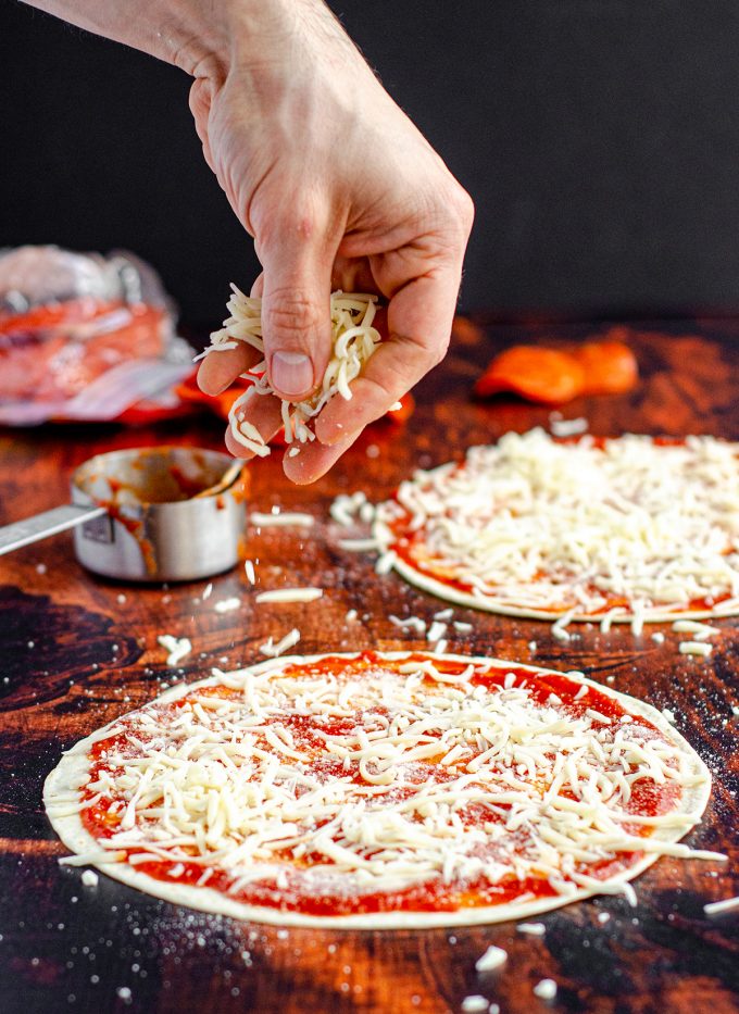 A hand sprinkling shredded mozzarella onto a tortilla to make tortilla pizza.