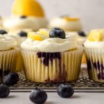 A lemon blueberry cupcake sitting on a metal grater with a blueberry on top and a lemon slice on top and beside it.