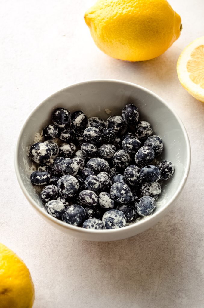 A bowl of blueberries that have been tossed in flour with lemons around it.