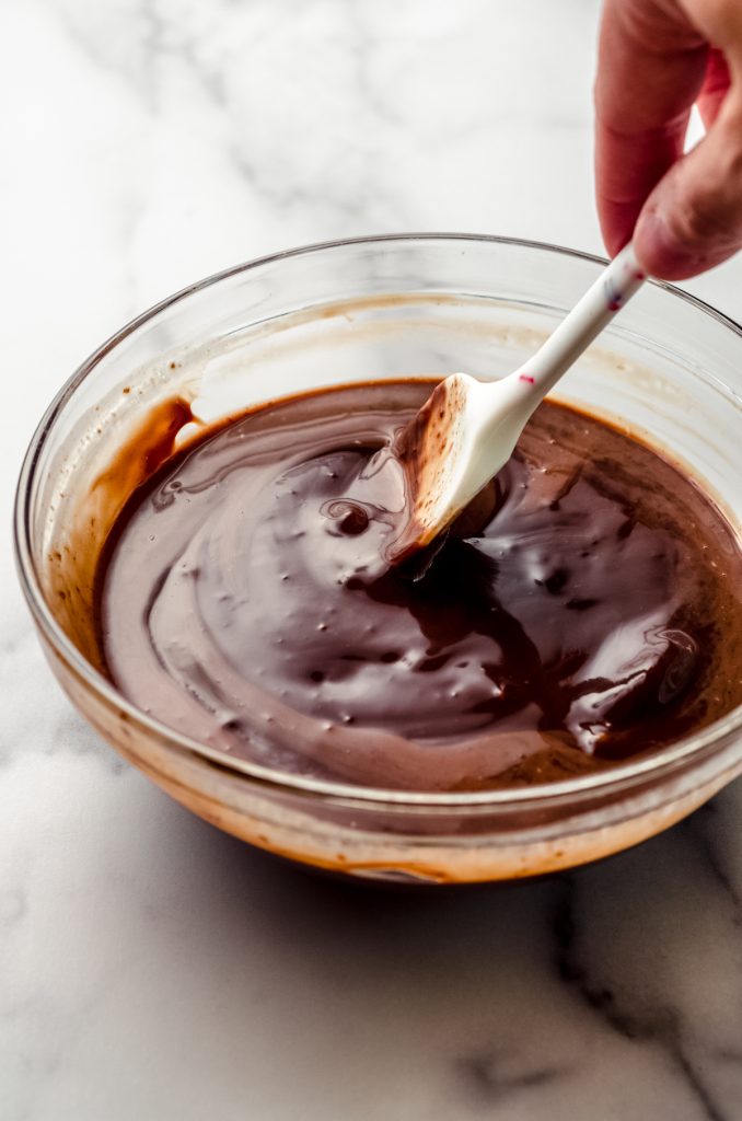 Someone is stirring chocolate ganache in a glass bowl with a spatula.