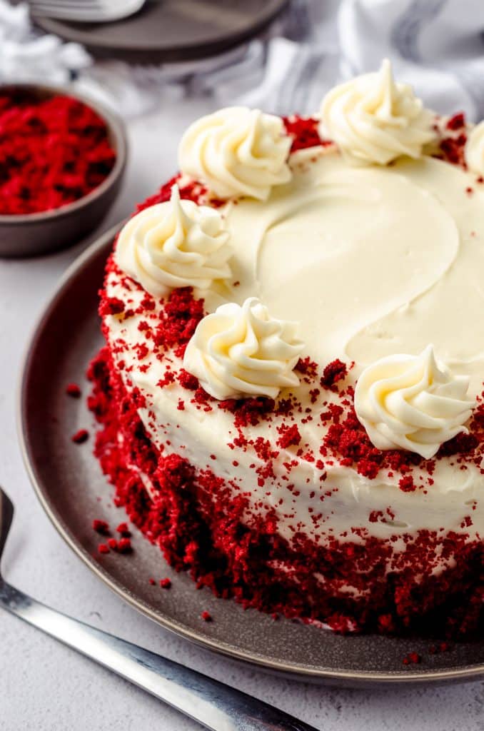 A red velvet cake with cream cheese frosting. There are red velvet cake crumbs and dollops of frosting on top.