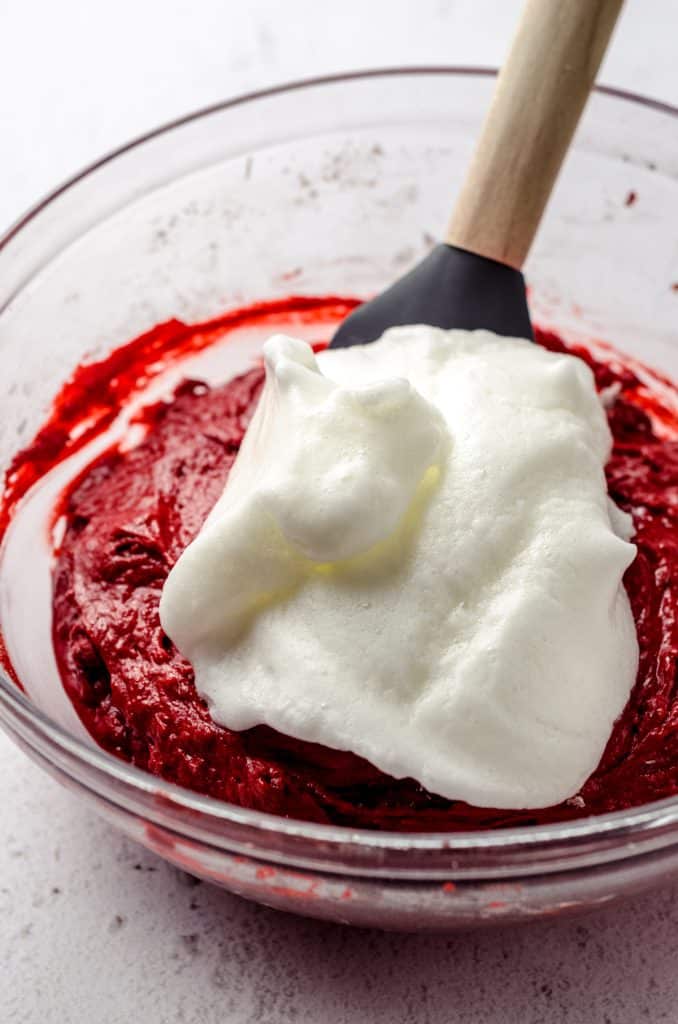 A pile of whipped egg whites on top of red velvet cupcake batter with a spatula ready to be folded into the batter.