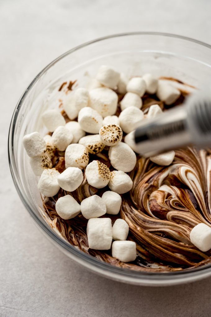 Someone is using a kitchen torch to toast marshmallows on top of a bowl of s'mores dip.
