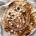 Aerial photo of a bowl of s'mores dip on a plate with mini pretzels and graham crackers scattered around the bowl.