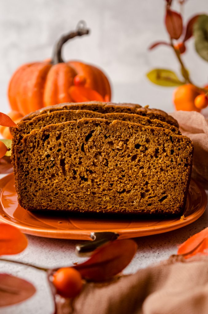 Slices of pumpkin bread loaf on a plate with pumpkins and fall decor in the background.