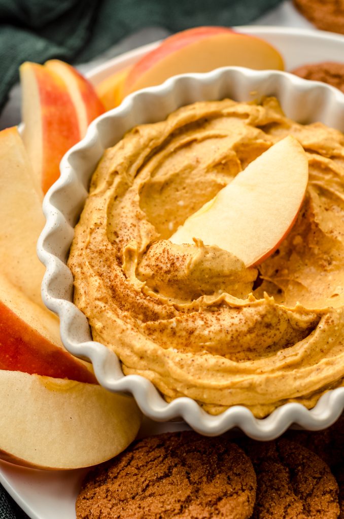 Pumpkin dip in a bowl on a plate with gingersnaps and apple slices. There is an apple slice in the bowl of dip.