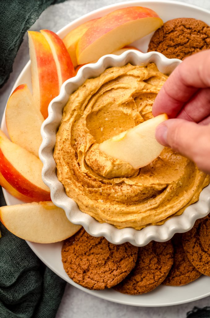 Someone is dipping an apple slice into a bowl of pumpkin dip.