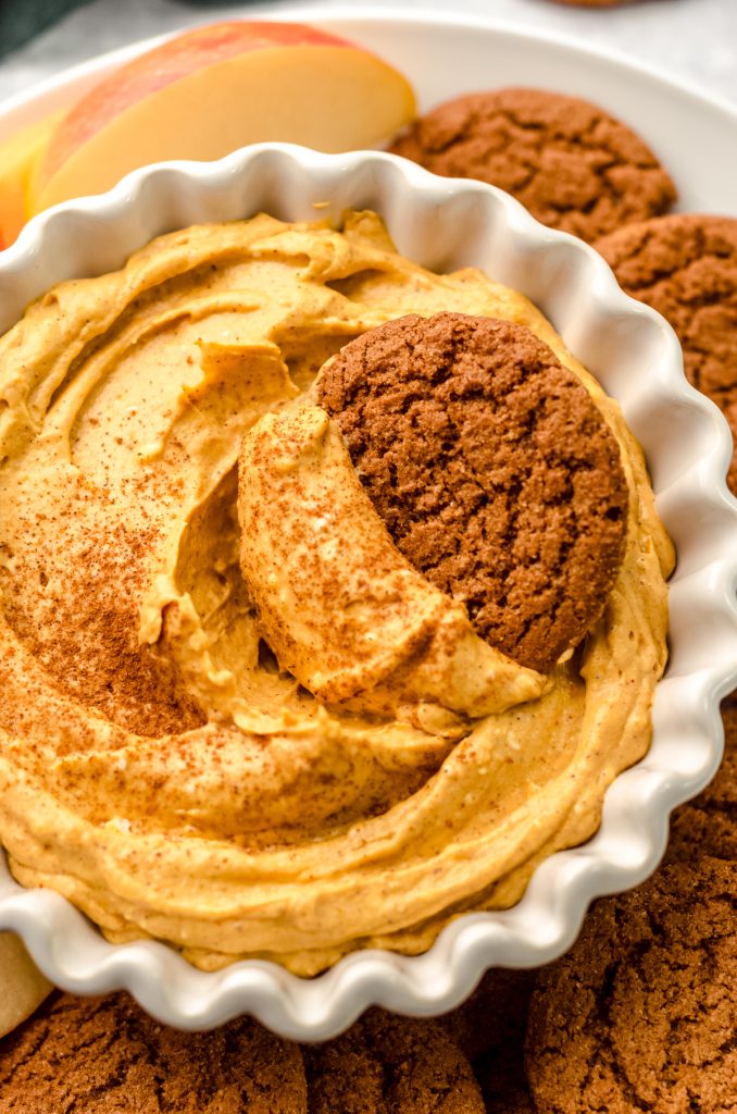 Pumpkin dip in a bowl on a plate with gingersnaps and apple slices. There is a gingersnap in the bowl of dip.