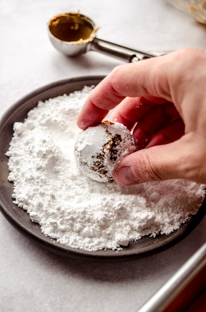 A ball of gingerbread crinkle cookie dough being rolled around in powdered sugar.