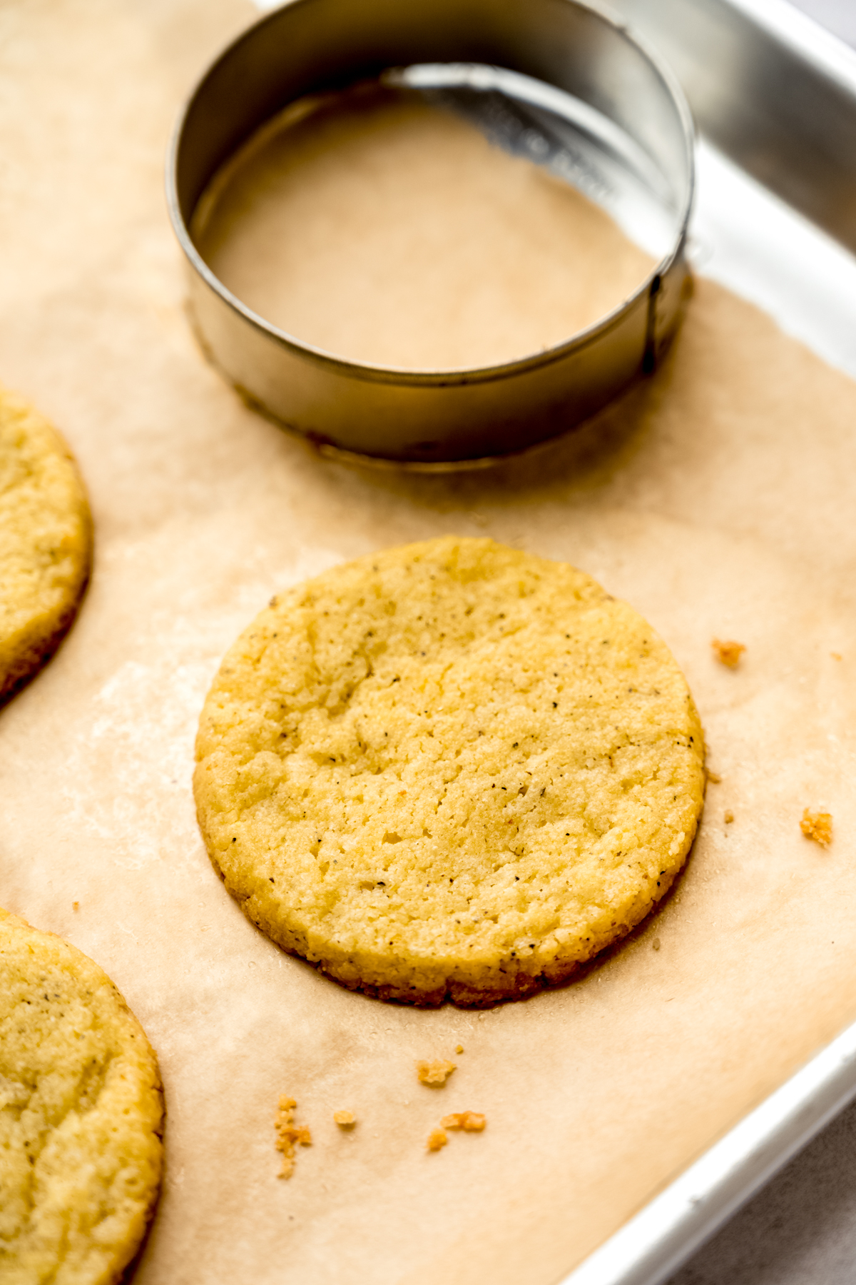 A lemon pepper cookie on a baking sheet with a circle cookie cutter off to the side that was used to reshape the hot cookie.