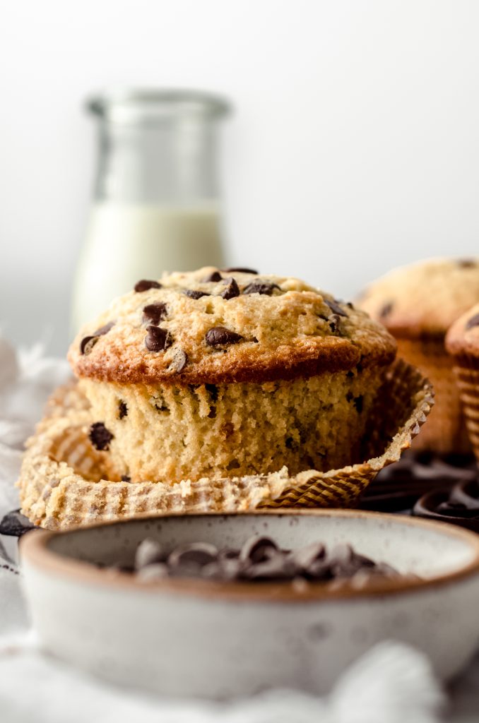 A jumbo chocolate chip muffin with the wrapper pulled down sitting on a cooling rack with a bottle of milk in the background.
