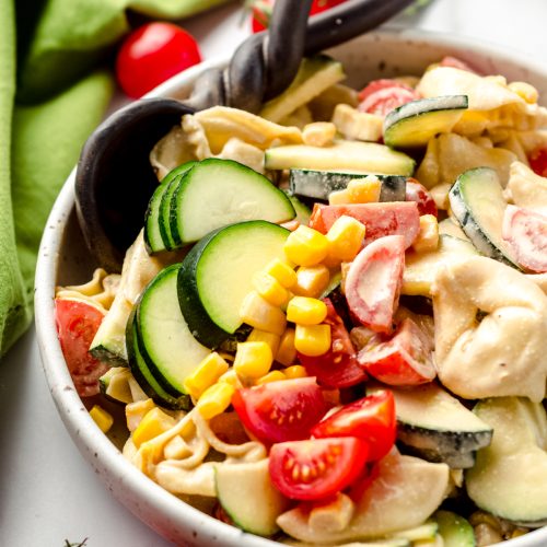 Tortellini pasta salad with zucchini, corn, and tomatoes in a bowl with a serving spoon.