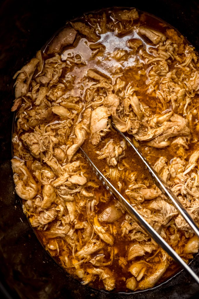 Aerial photo of shredded hot chicken in a slow cooker with tongs in it.