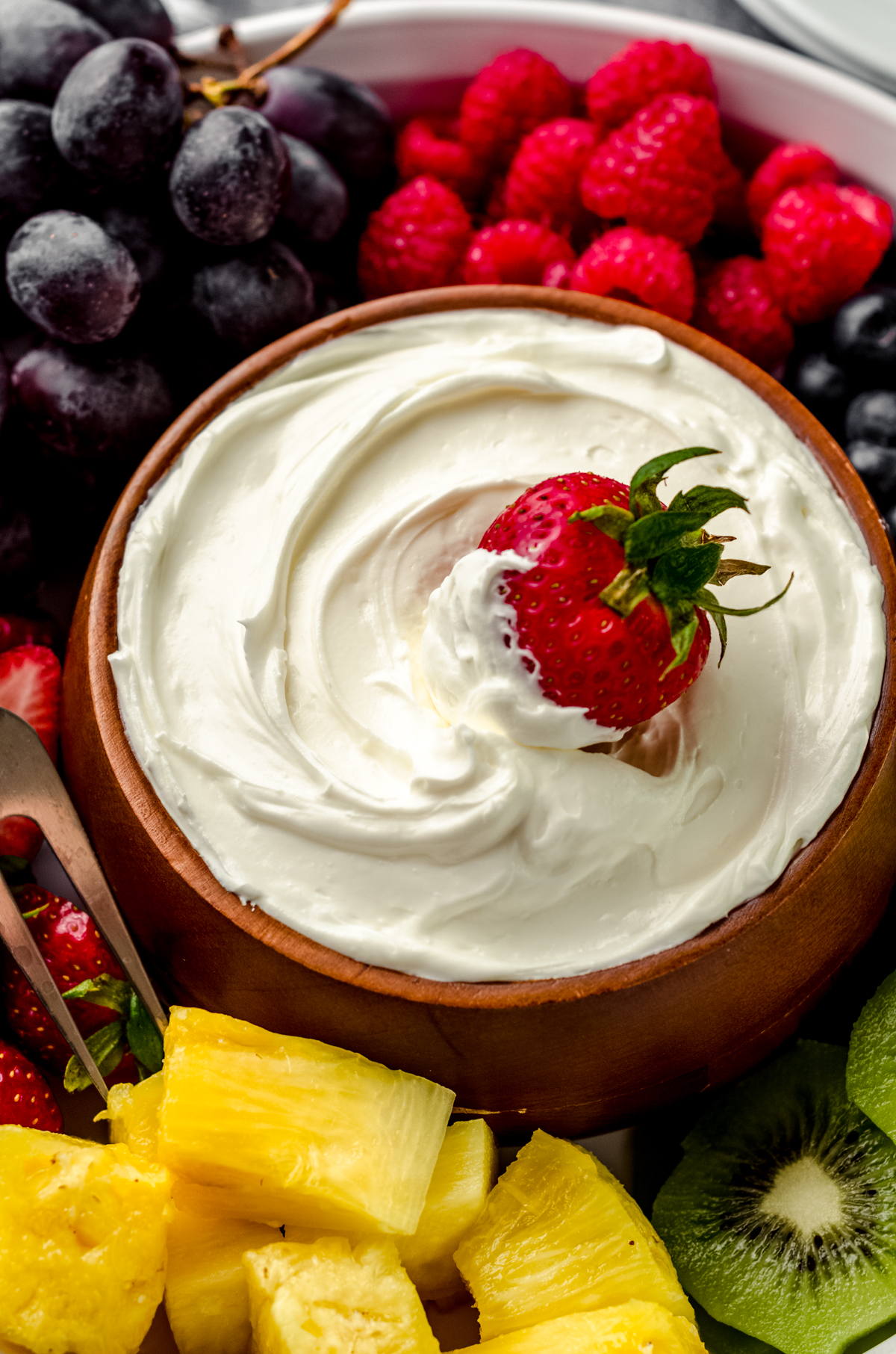 A bowl of fruit dip with a strawberry in it surrounded by a variety of sliced fruits.