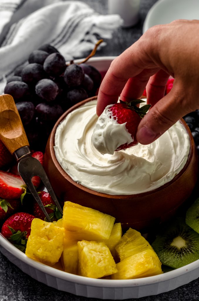 A bowl of fruit dip surrounded by a variety of sliced fruits. A hand is dipping a strawberry into the dip.