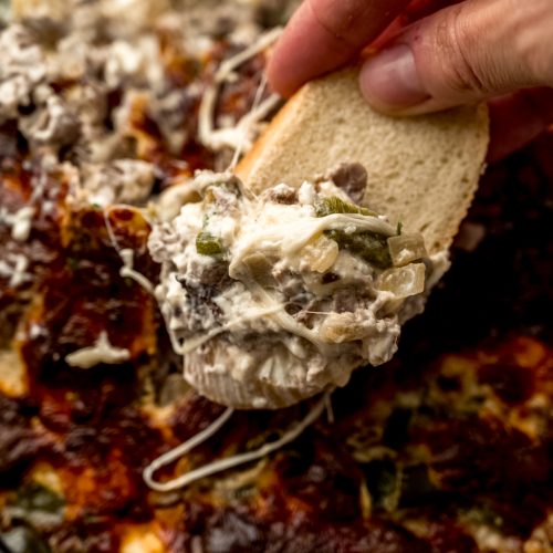 Someone holding a baguette piece that has been dipped into Philly cheesesteak dip.