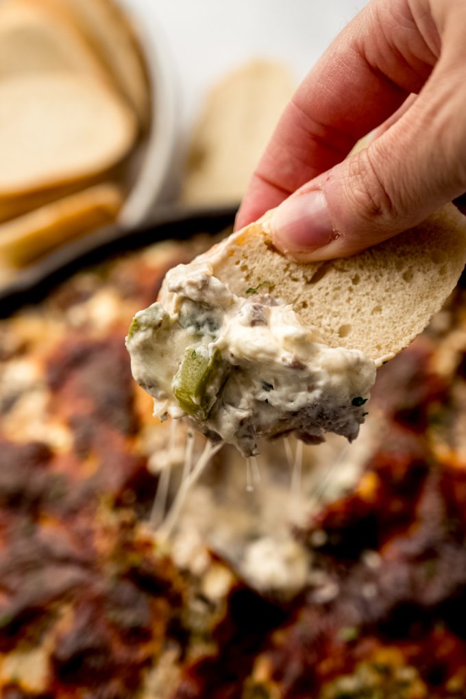 Someone holding a baguette piece that has been dipped into Philly cheesesteak dip.