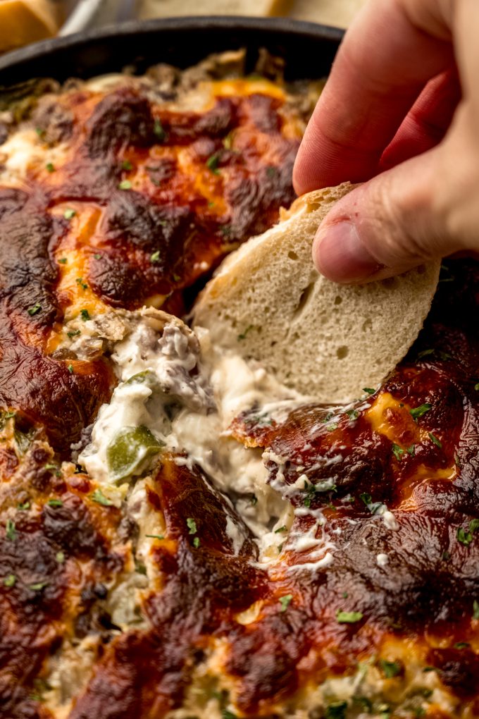 Someone is dipping a baguette piece into Philly cheesesteak dip.