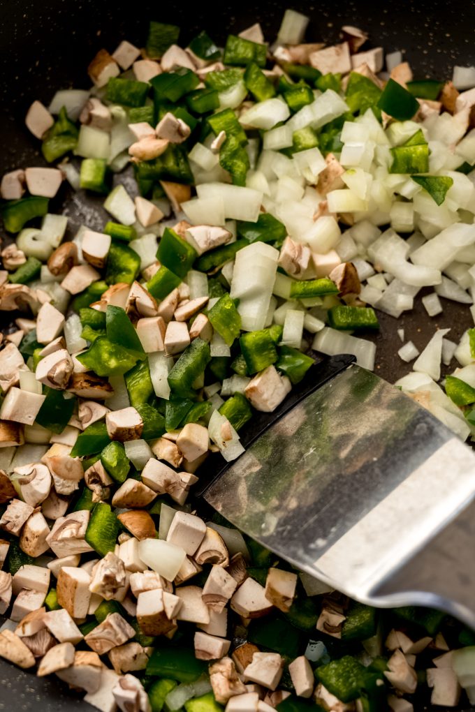 Onions, peppers, and mushrooms cooking in a pan with a spatula.
