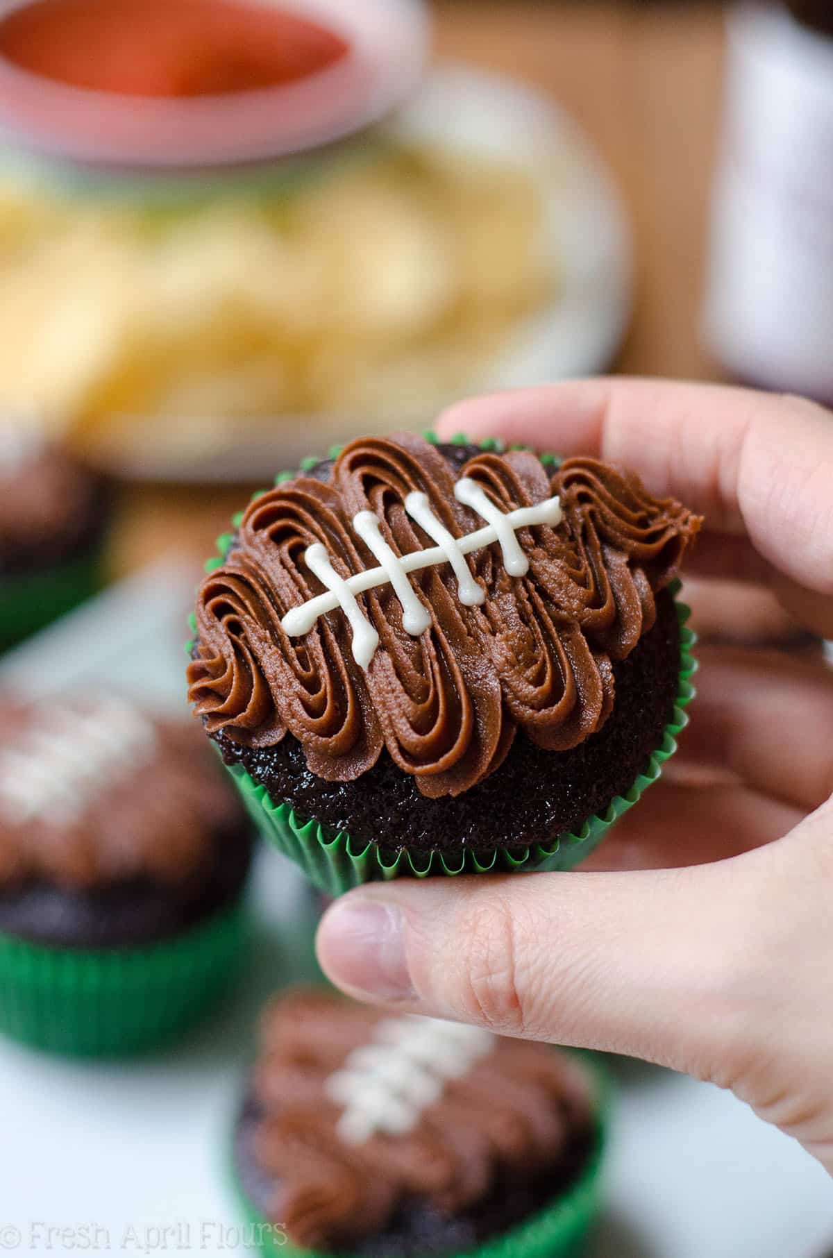 a hand holding a chocolate cupcake in green cupcake liner with a chocolate frosting football piped on top to make a football cupcake