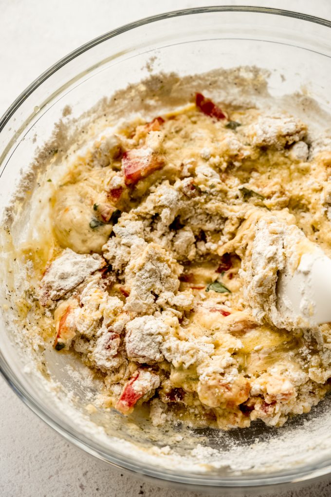 A large glass bowl with tomato basil bread batter and a spatula in it.