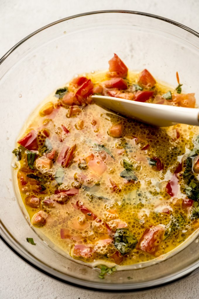 A large glass bowl with tomato basil bread batter and a spatula in it.