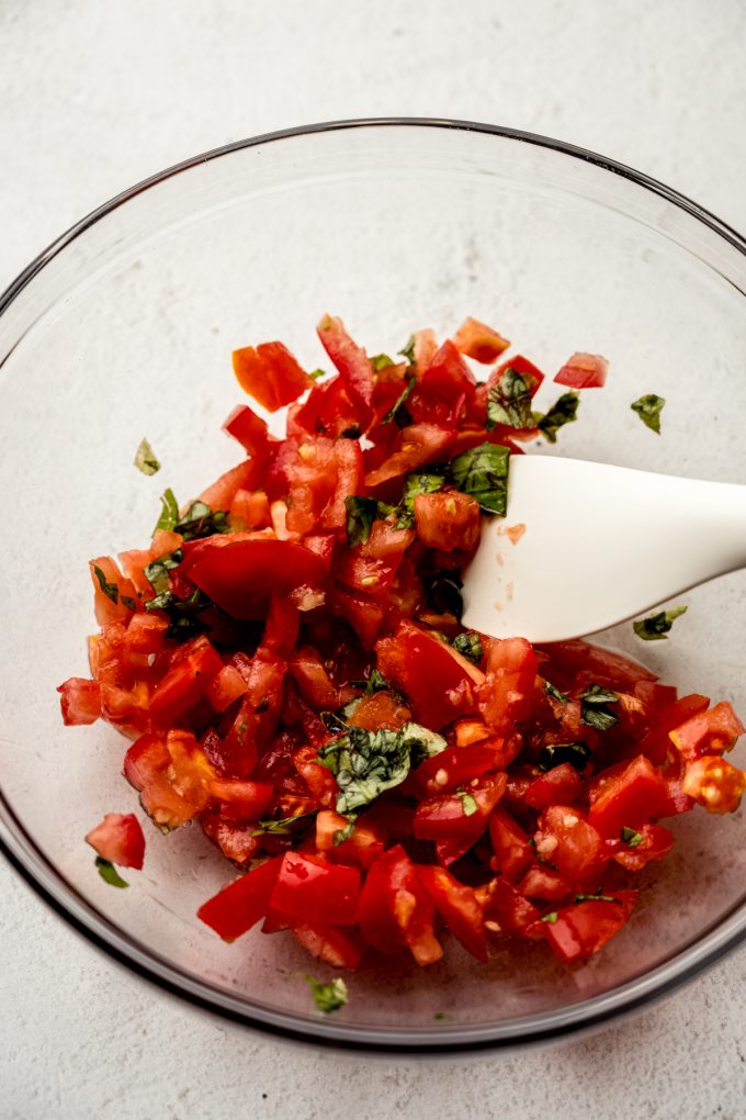Diced tomatoes and basil getting tossed together in a large glass bowl with a spatula.