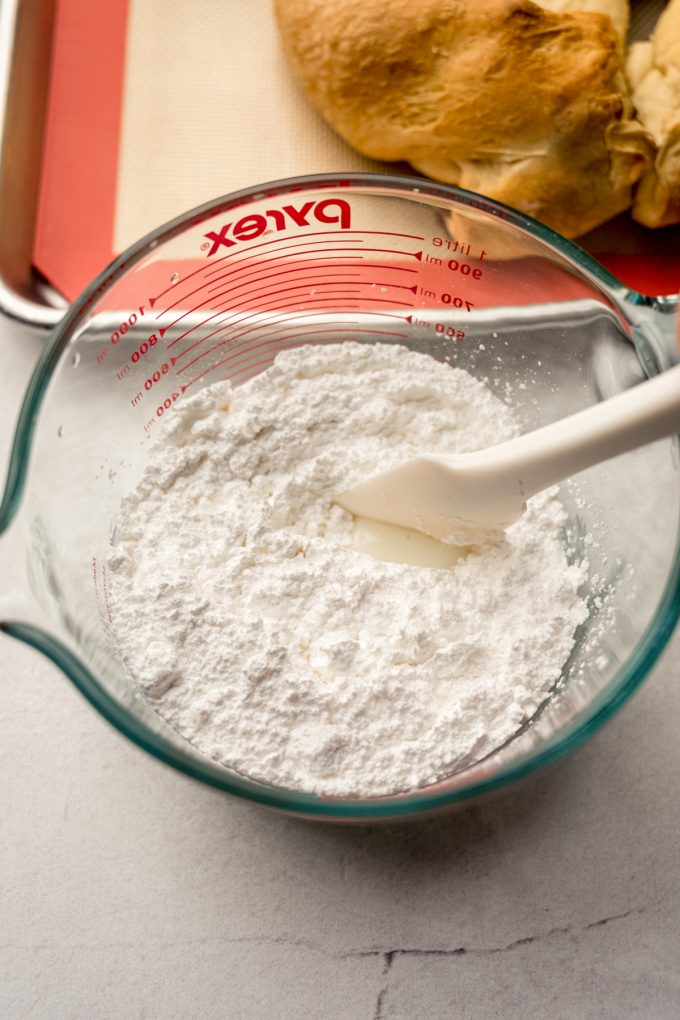 Someone is stirring icing with a spatula in a glass measuring cup.