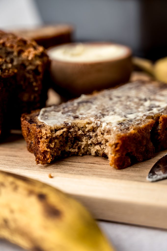 A slice of buttered banana nut bread on a cutting board with a bite taken out of it.