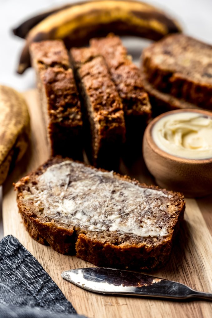 A slice of buttered banana nut bread on a cutting board other slices and a bowl of butter behind it.