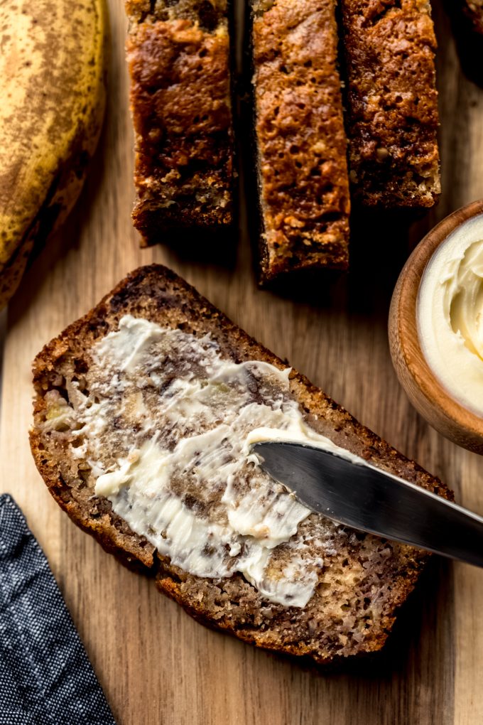 Aerial photo of someone using a butter knife to spread butter onto a slice of banana nut bread.