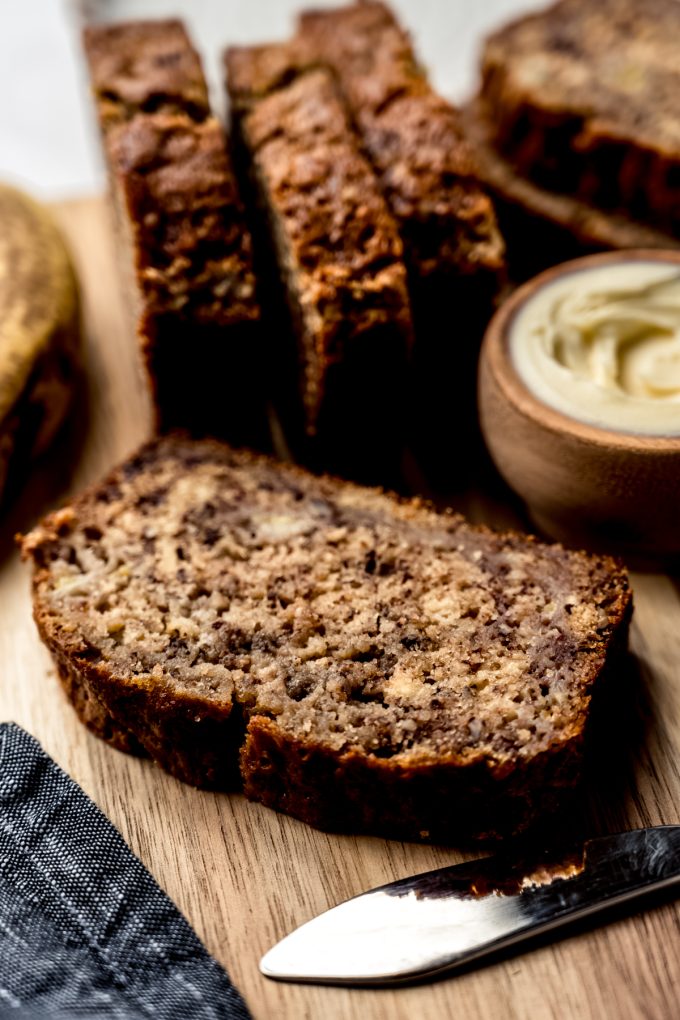 A slice of banana nut bread on a cutting board other slices and a bowl of butter behind it.