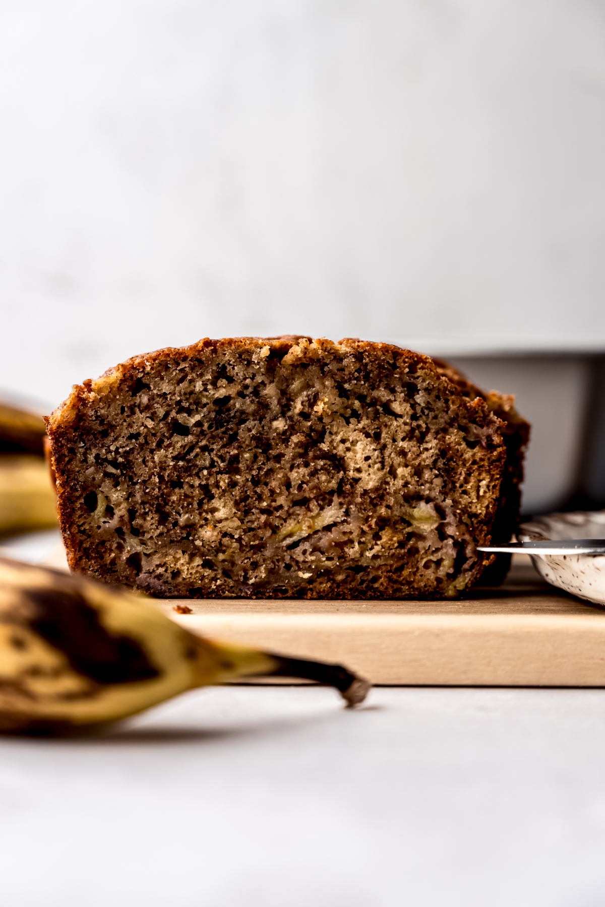 A loaf of banana bread on a cutting board with a banana in the foreground and a loaf pan and more bananas in the background.