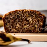 A loaf of banana bread on a cutting board with a banana in the foreground and a loaf pan and more bananas in the background.