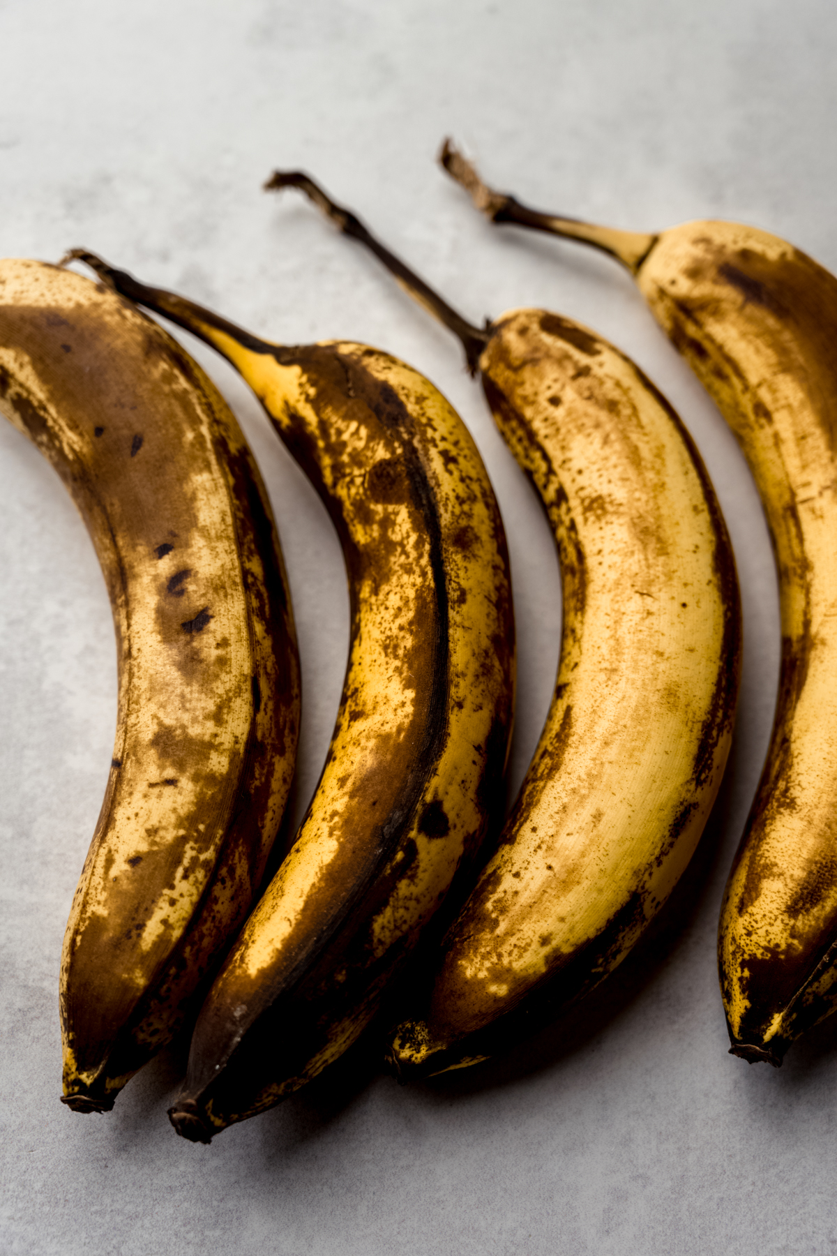 Four ripe bananas with lots of brown spots on them on a surface.