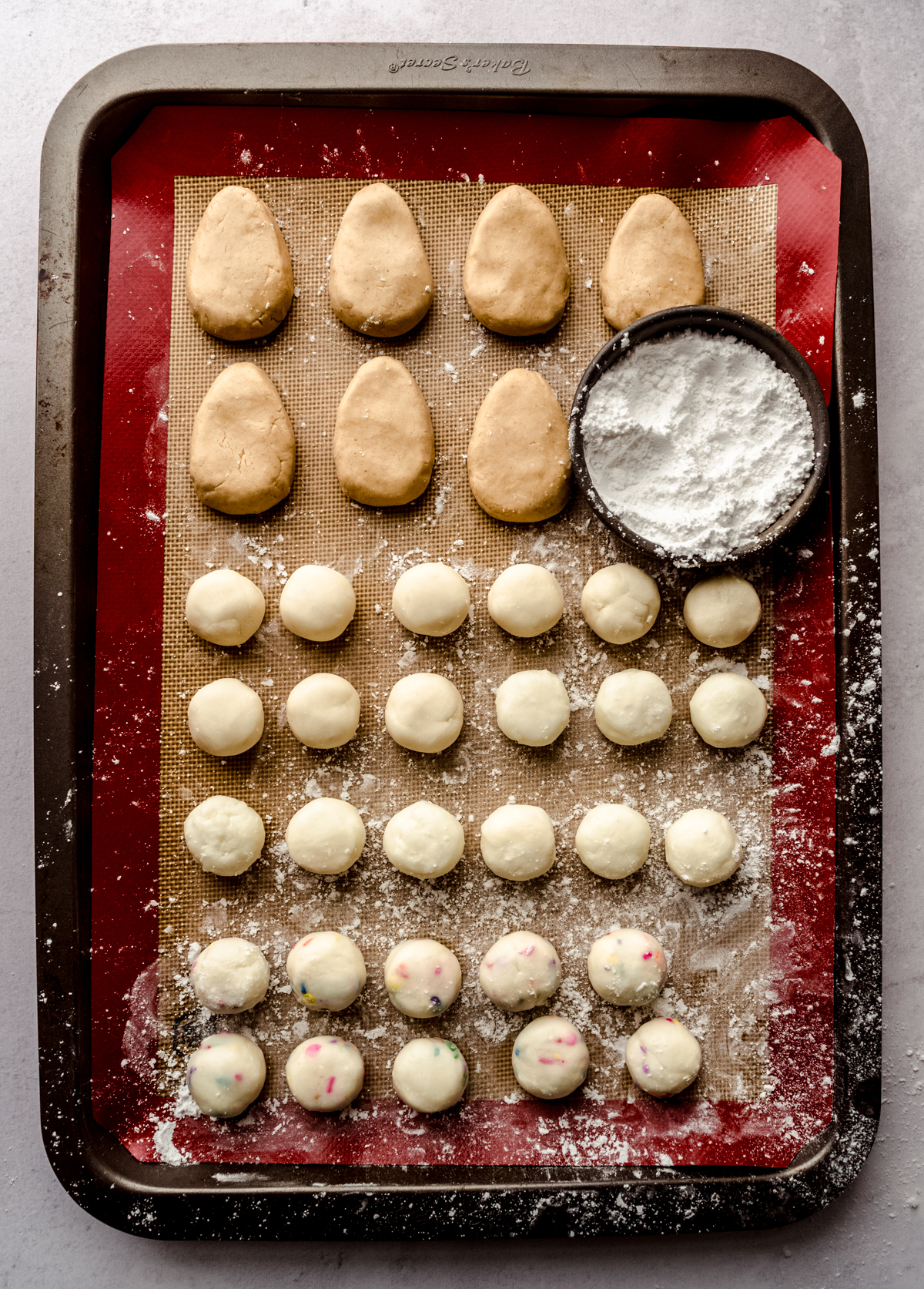 Aerial photo of buttercream Easter eggs and rounds on a baking sheet with a bowl of powdered sugar.