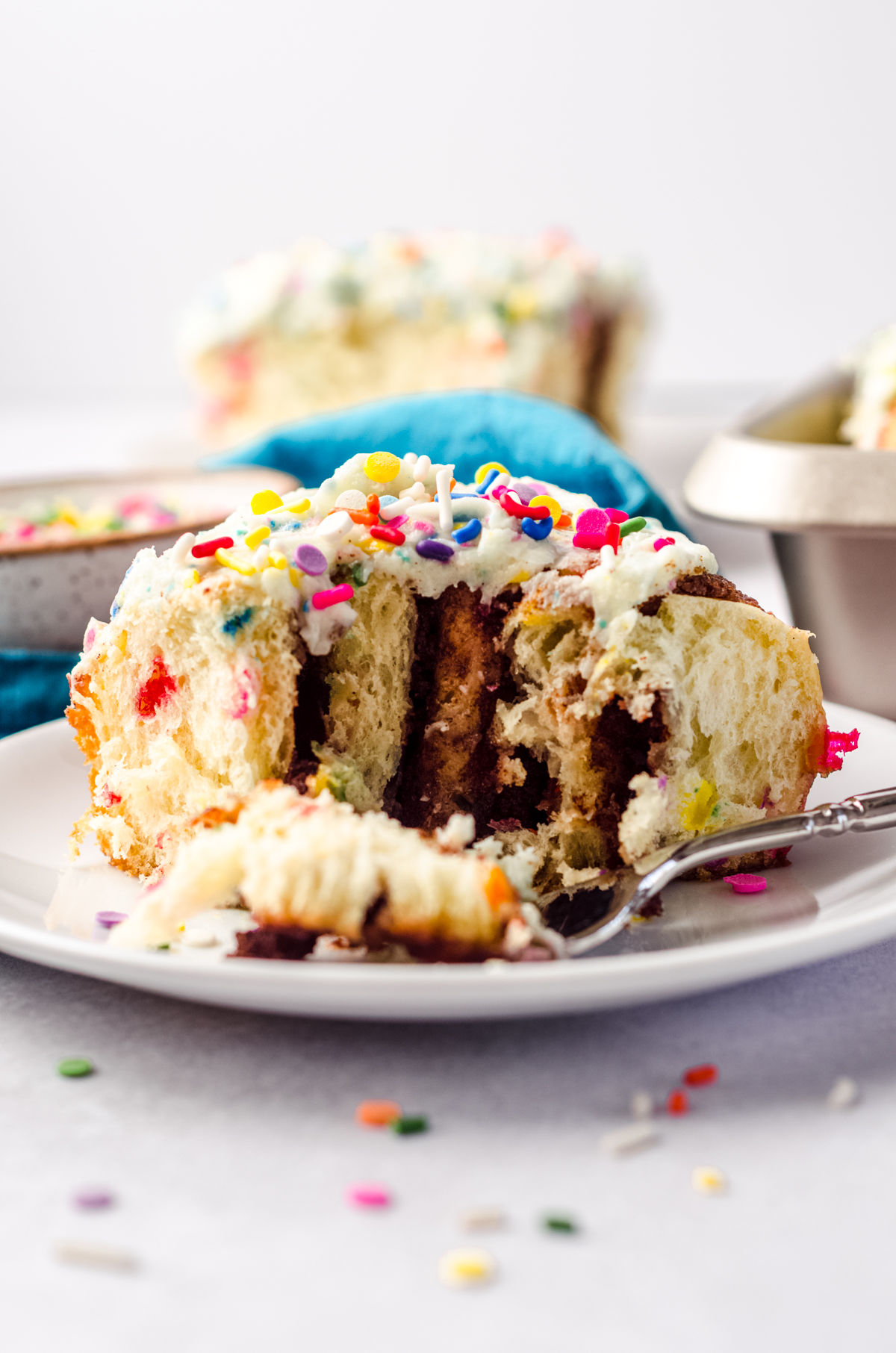 A funfetti cinnamon roll on a plate with a fork holding a bite of the roll.