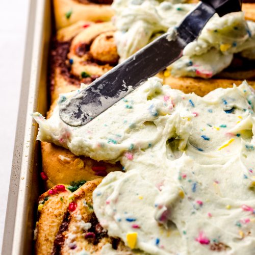 Someone is using a spatula to spread homemade rainbow chip frosting onto funfetti cinnamon rolls.