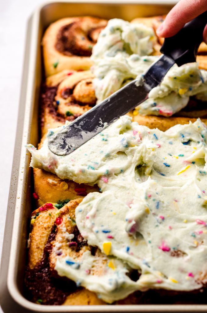Someone is using a spatula to spread homemade rainbow chip frosting onto funfetti cinnamon rolls.