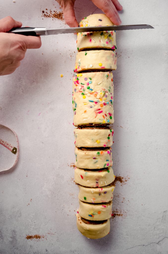 Someone is slicing a funfetti cinnamon roll log with a knife.