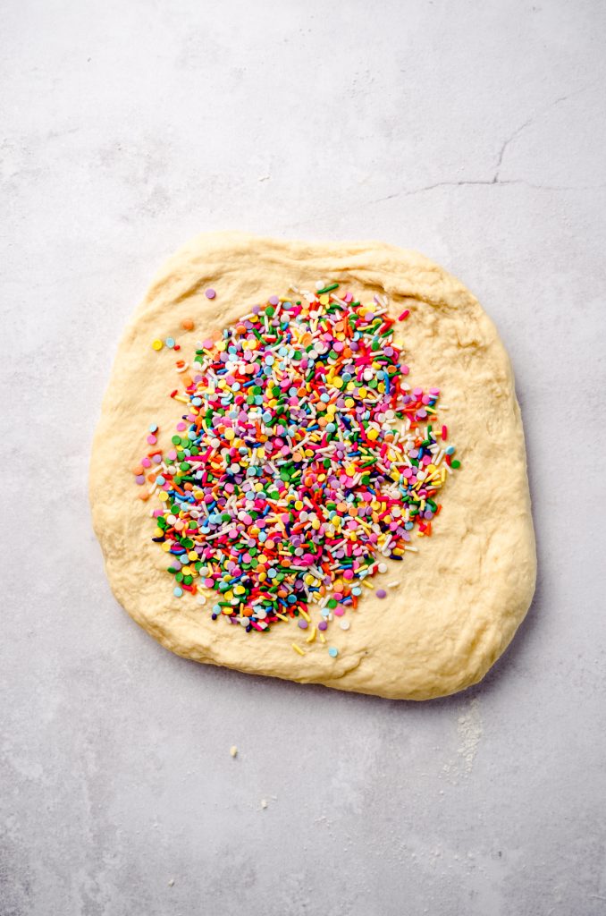 A slab of cinnamon roll dough with sprinkles on it ready to be kneaded into the dough.