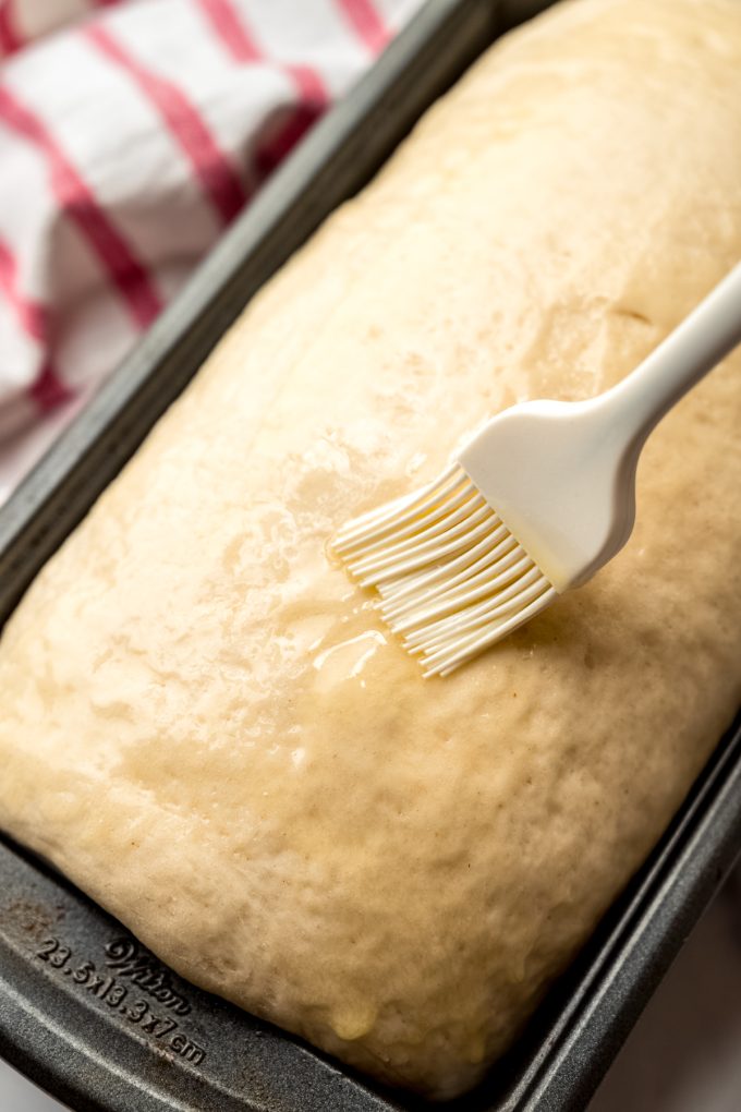 Someone is brushing melted butter onto a log of sandwich bread dough in a loaf pan.