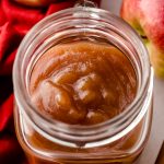 A jar of homemade apple butter with apples and a bowl of apple butter around it.