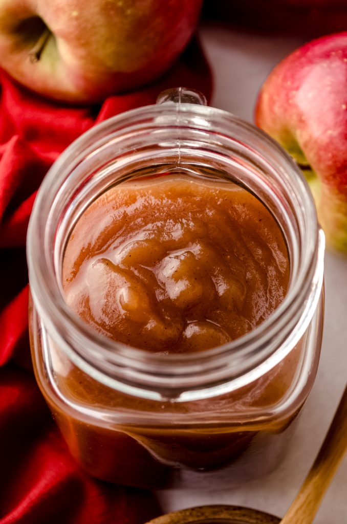 A jar of homemade apple butter with apples and a bowl of apple butter around it.