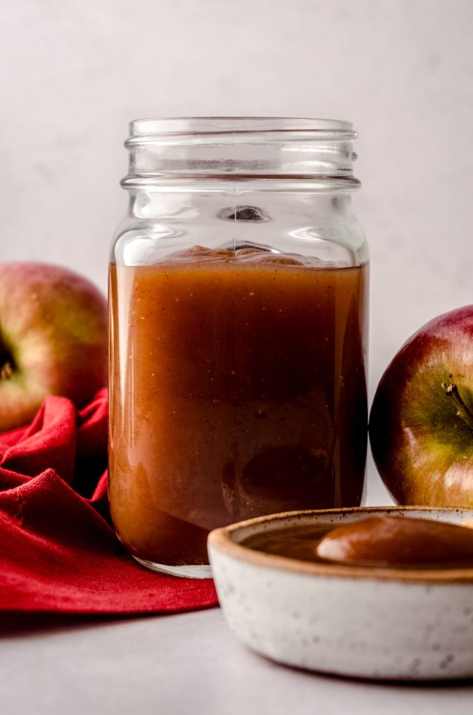 A jar of homemade apple butter with apples and a bowl of apple butter around it.