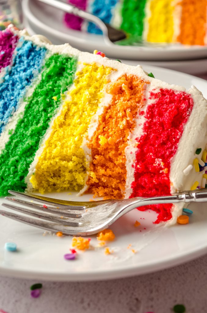 A closeup of a slice of rainbow cake on a plate with a fork and a bite has been taken out of the cake.