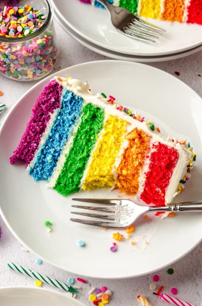 A slice of rainbow cake on a plate with a fork and a bite has been taken out of the cake.