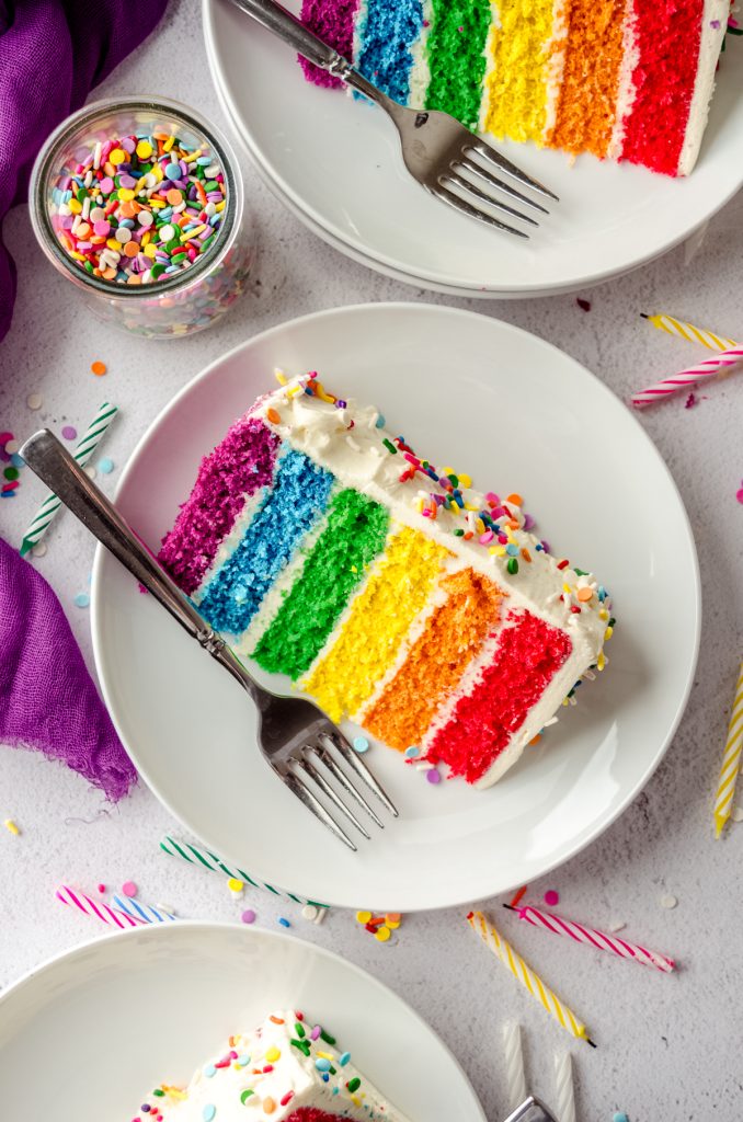 Aerial photo of a slice of rainbow layer cake on a plate with a fork and sprinkles around it.