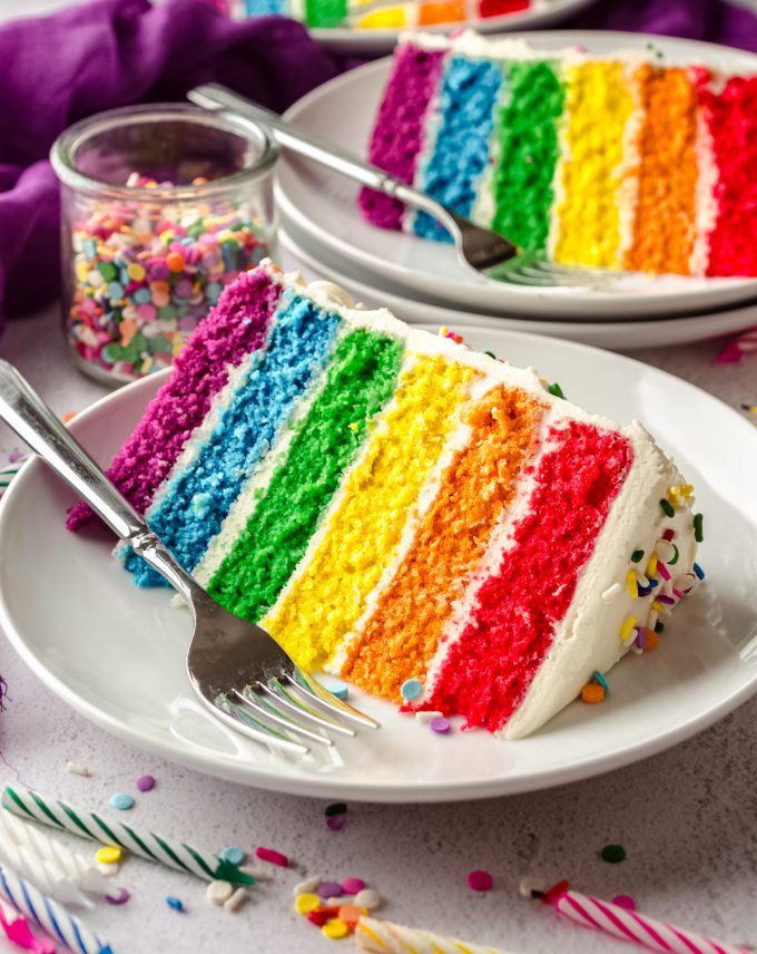 A slice of rainbow layer cake on a plate with a fork and sprinkles around it.