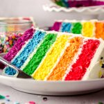 A slice of rainbow layer cake on a plate with a fork and sprinkles around it.